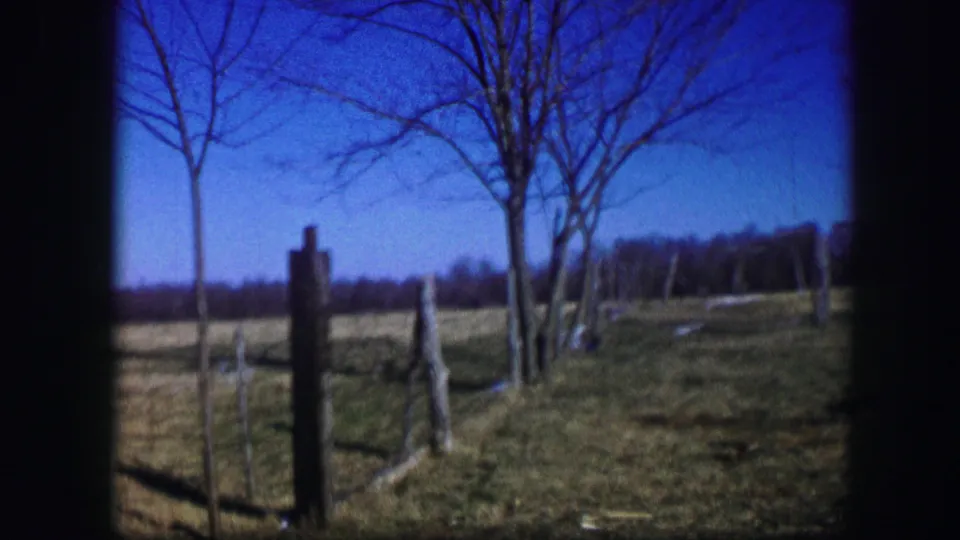 1967 Kirksville rural frame showing fence posts, bare trees, open grass, distant tree line, and a clear blue sky.