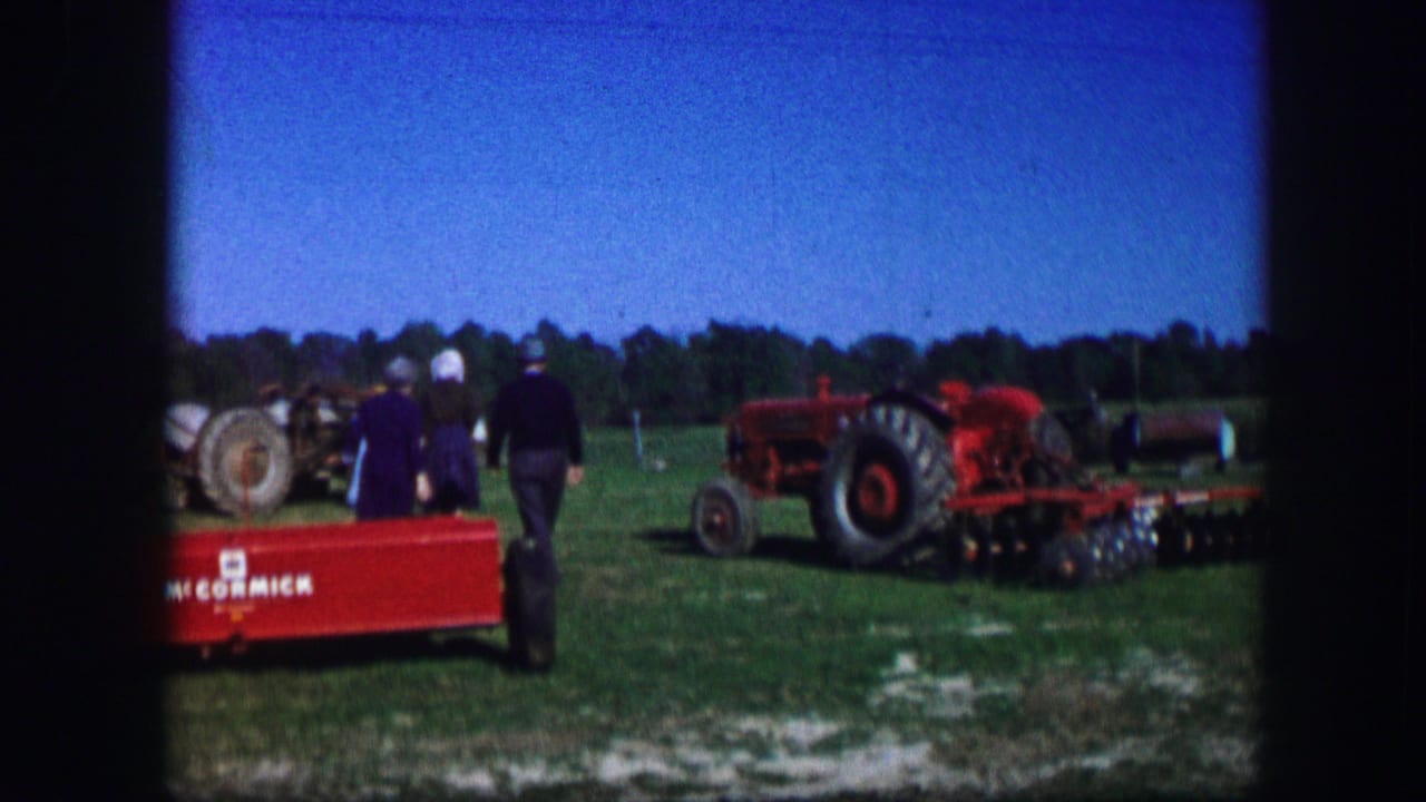 Adults stand near red farm equipment and a tractor on a grassy fairground.