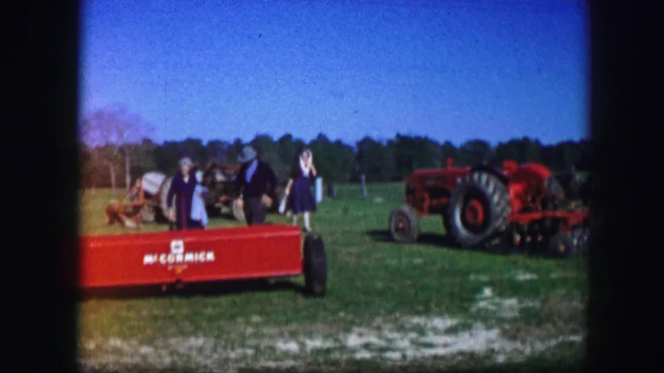 Home-movie frame from 1967 Kirksville showing fairgoers near red farm machinery and a tractor on grass.