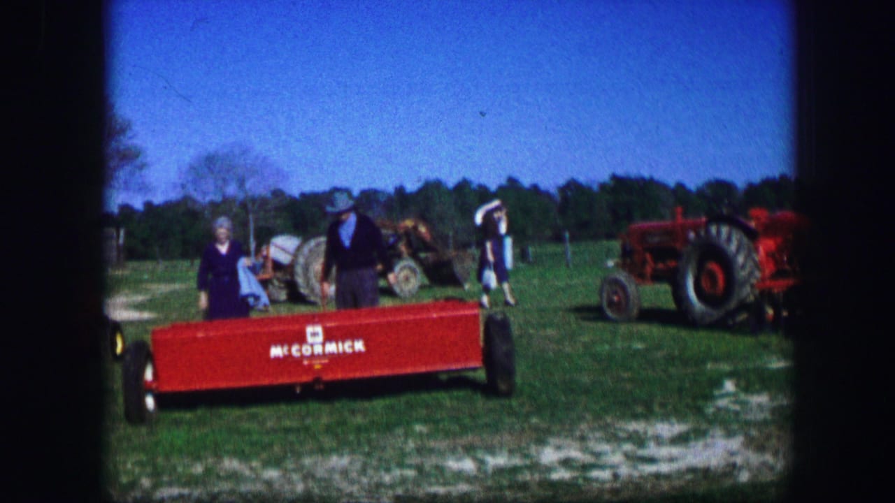 1967 Kirksville, Missouri home-movie frame showing visitors beside a red McCormick farm attachment with tractors behind them.