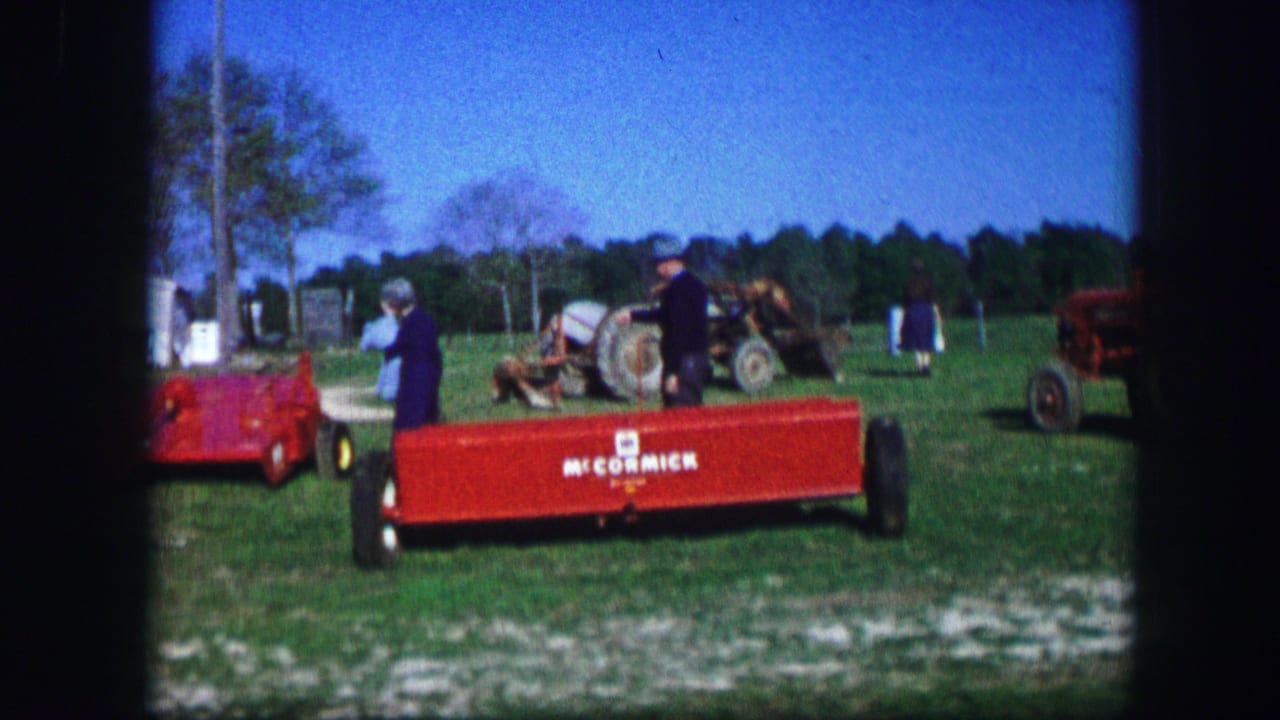 Red McCormick-branded trailer in a grassy field with adults, white trailers, and farm equipment behind it.
