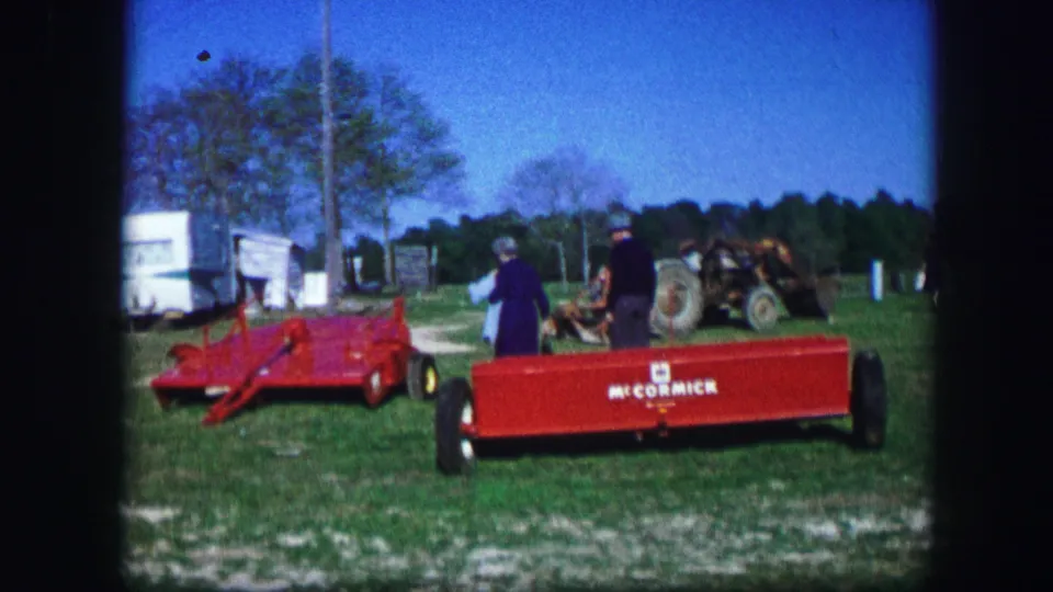 1967 Kirksville, Missouri home-movie frame showing red equipment, a McCormick-marked trailer, adults, white trailers, and a farm machine in a rural field.