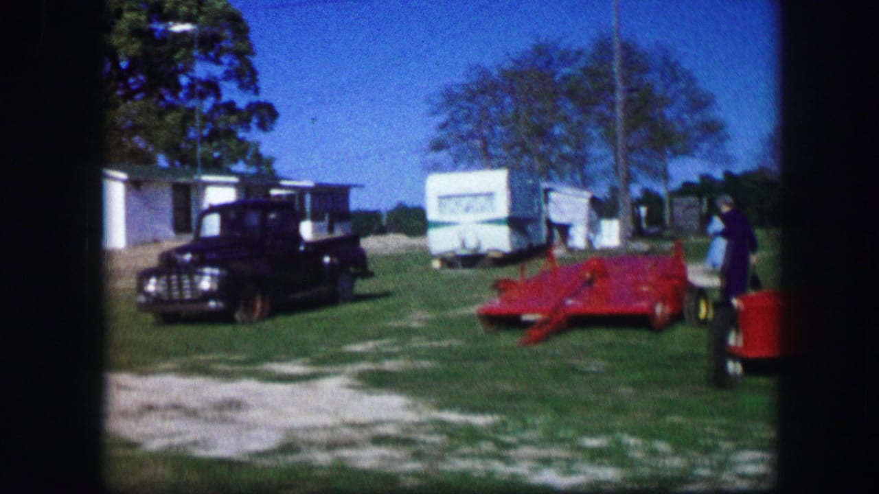 Black truck and red farm implement beside trailers on a grassy rural campsite.