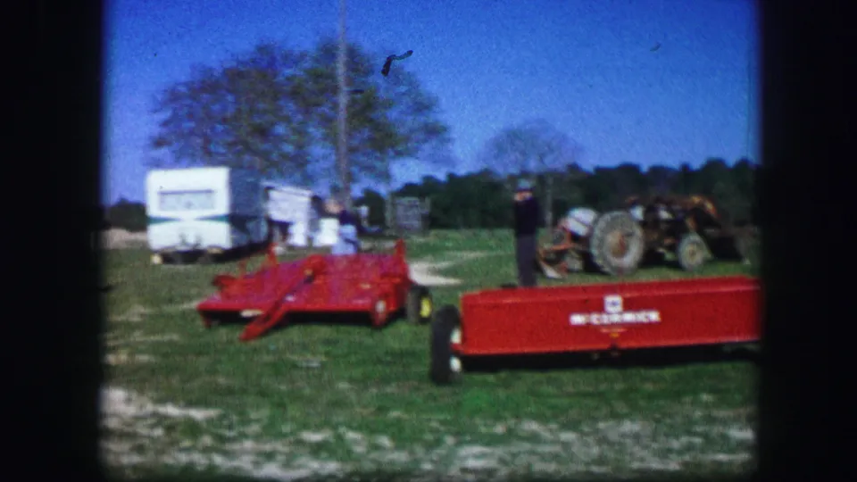 1967 rural Missouri campsite scene with white trailers, a red wagon or implement, tractor, grassy ground, and an adult near the equipment.
