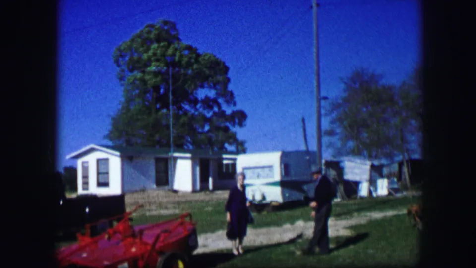 At a Kirksville, Missouri campsite, two adults stand near a red farm implement, small white building, white trailer, trees, and grassy ground.