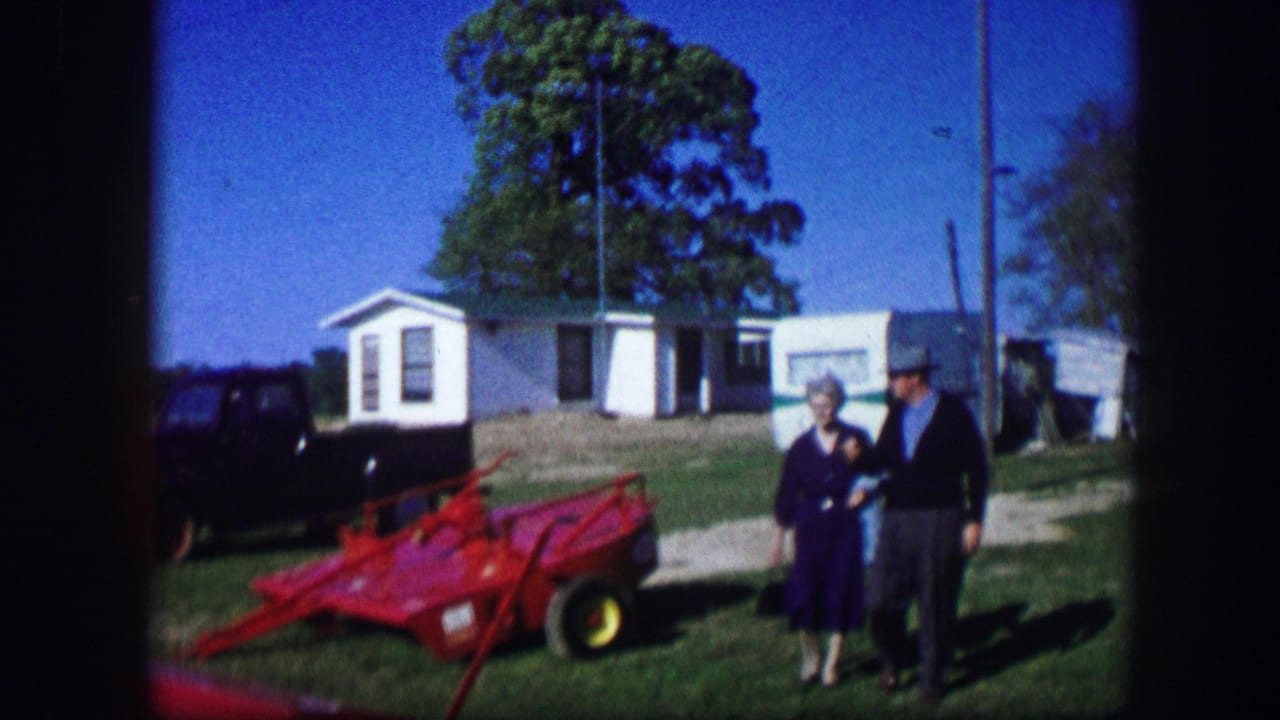 In Kirksville, Missouri, 1967, a woman and man stand near red farm equipment with small buildings, a dark car, and trees behind them.