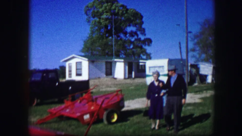 In Kirksville, Missouri, 1967, a woman and man stand near red farm equipment with small buildings, a dark car, and trees behind them.