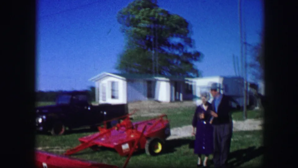 The 1967 Kirksville home movie shows two adults near a red farm implement, with a dark car and white outbuildings or trailers in the background.