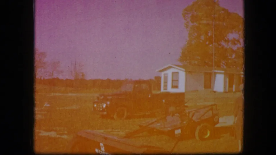 A more overexposed 8mm frame from the Kirksville scene shows the red farm implement, yard, distant vehicle, trees, and small white building.