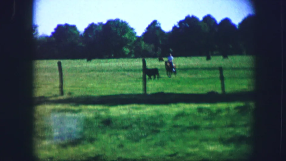 Wide 1967 Kirksville pasture frame showing a rider near a dark animal beyond a fence, with trees across the background.