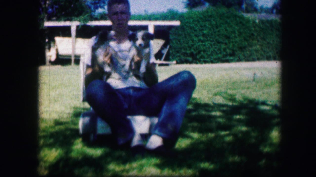 In 1967 Kirksville, Missouri home-movie footage, a man sits on a lawn chair holding two small puppies in a green yard.