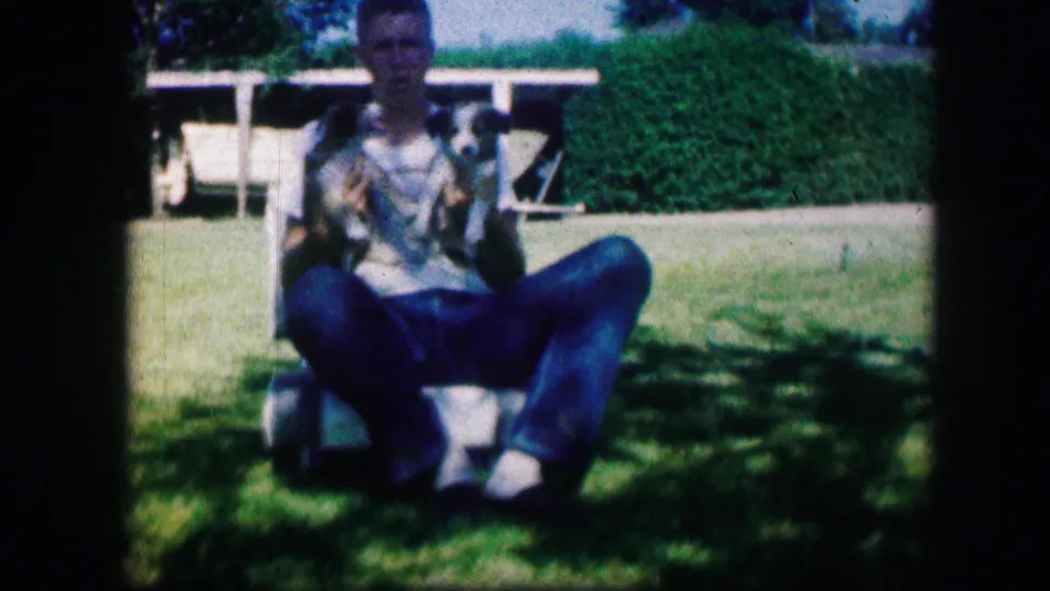 In 1967 Kirksville, Missouri home-movie footage, a man sits on a lawn chair holding two small puppies in a green yard.