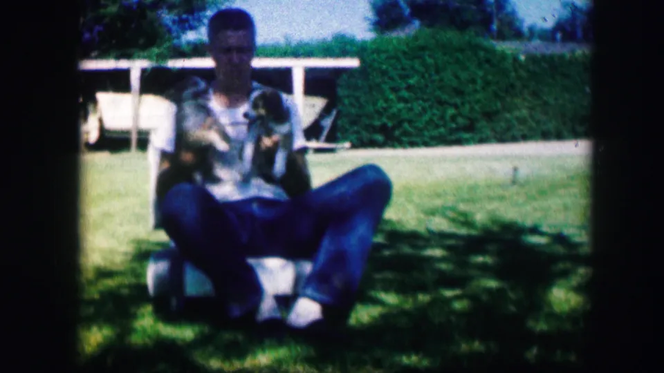 The man remains seated outdoors in the Kirksville yard, holding the puppies as they face outward against a hedge and white fence.