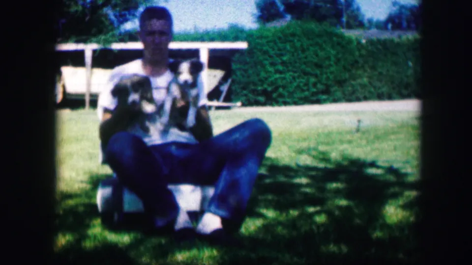 A 1967 home-movie frame shows the seated man holding two puppies close together in a shaded patch of the grassy yard.