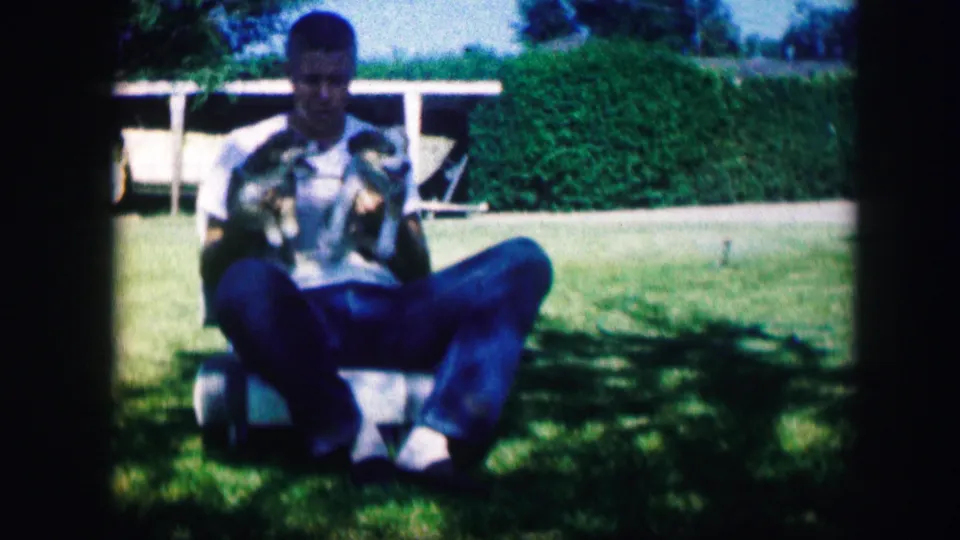 Near the end of the clip, the man sits in the Kirksville yard with both puppies in his lap, framed by grass, hedge, and a white rail.