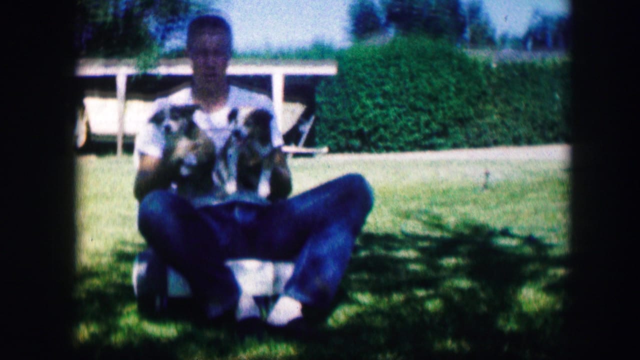 In 1967 Kirksville, Missouri home-movie footage, a young man in a white T-shirt and jeans sits on grass holding two puppies.