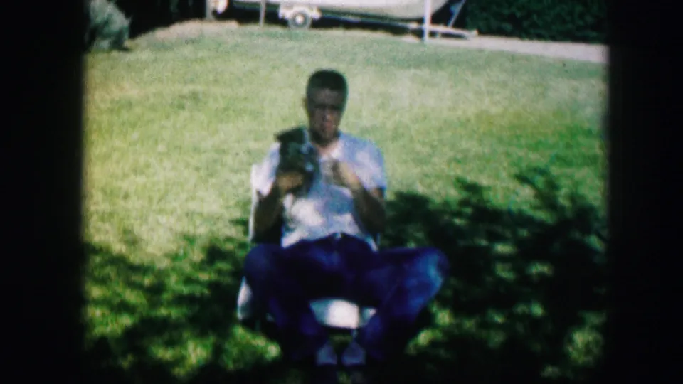 A young man in 1967 Kirksville backyard footage sits on a lawn chair and handles a small puppy in his lap.