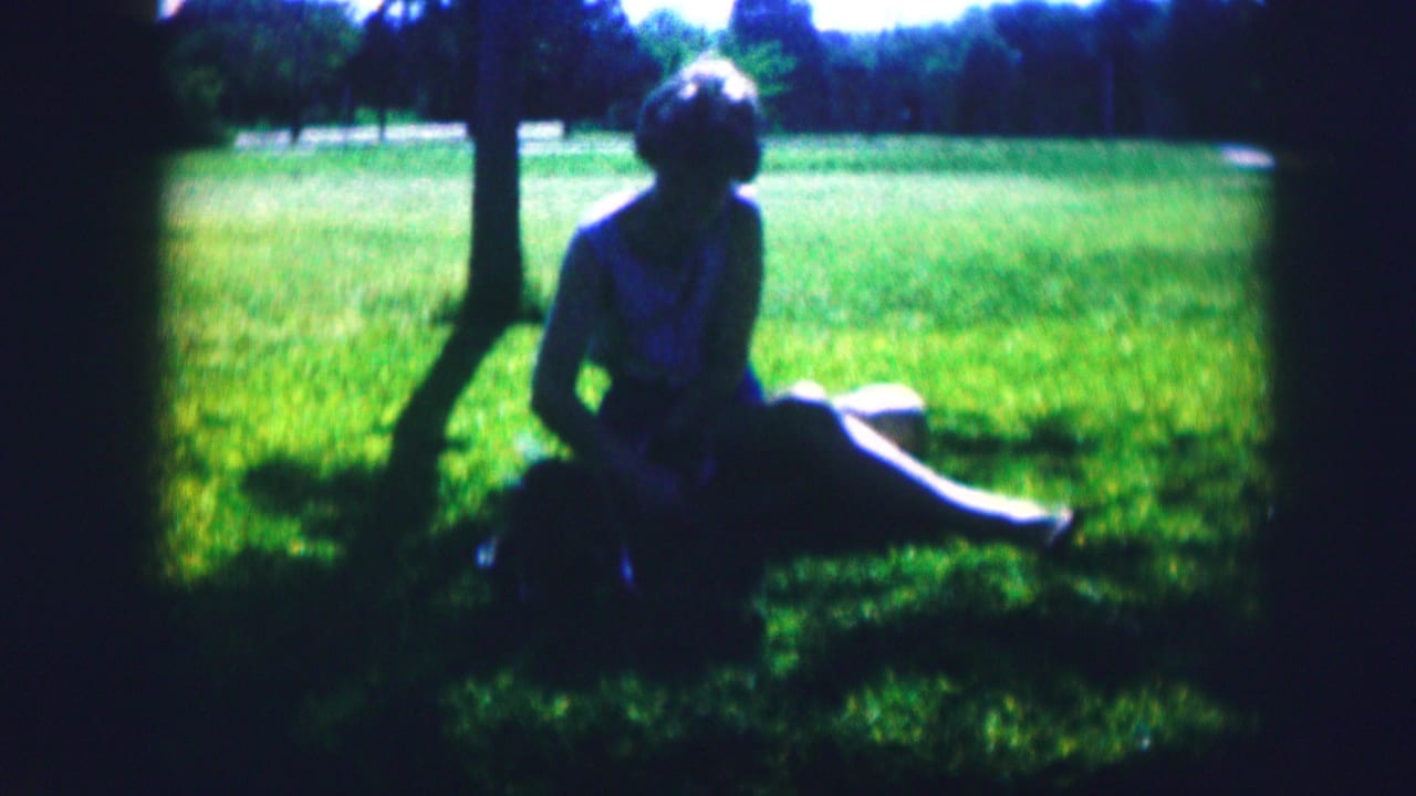 1967 Kirksville park frame showing a woman in a light dress seated in grass under shade with a small dog nearby.