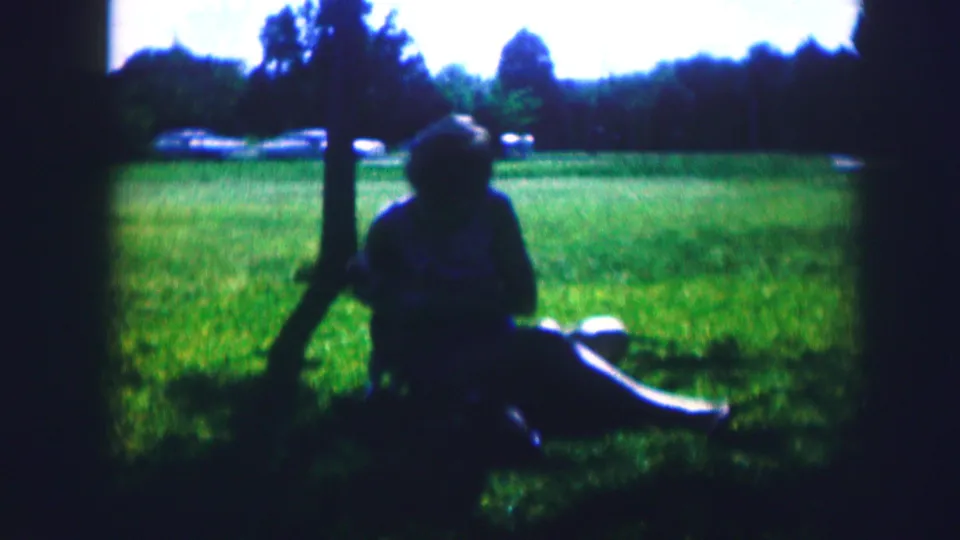 1967 Kirksville, Missouri 8mm frame showing a woman seated in shade under a tree with a dog at her side.
