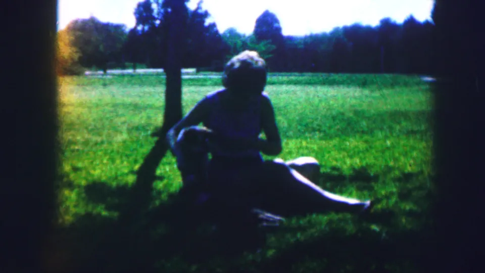 1967 Kirksville, Missouri 8mm frame showing a woman under a shade tree with a dog near her knees in a green field.