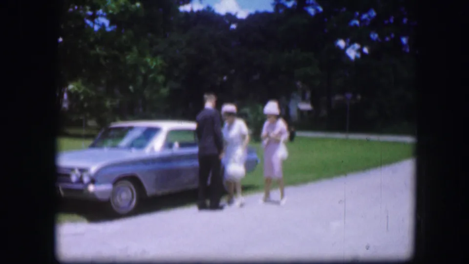 A suited man and two women in pale dresses are seen near a light blue car in the 1967 Kirksville outdoor home-movie scene.