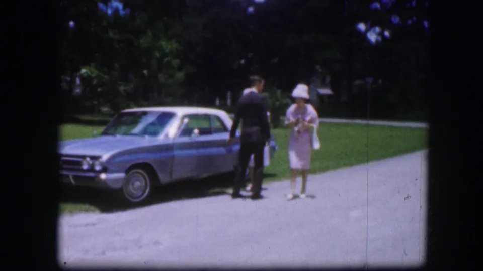 A woman in a pale dress and hat stands on the driveway as a suited man faces the light blue car in 1967 Kirksville footage.