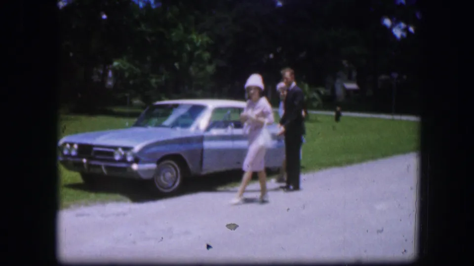 A woman in pale clothing walks in front of a light blue car while a suited man remains nearby in the Kirksville 1967 home movie.