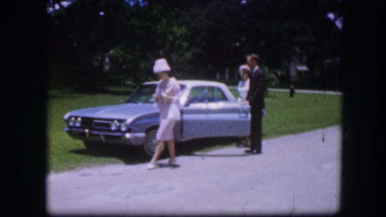 In 1967 Kirksville home-movie footage, a woman in a white hat stands near the front of a silver sedan with two adults beside the car.