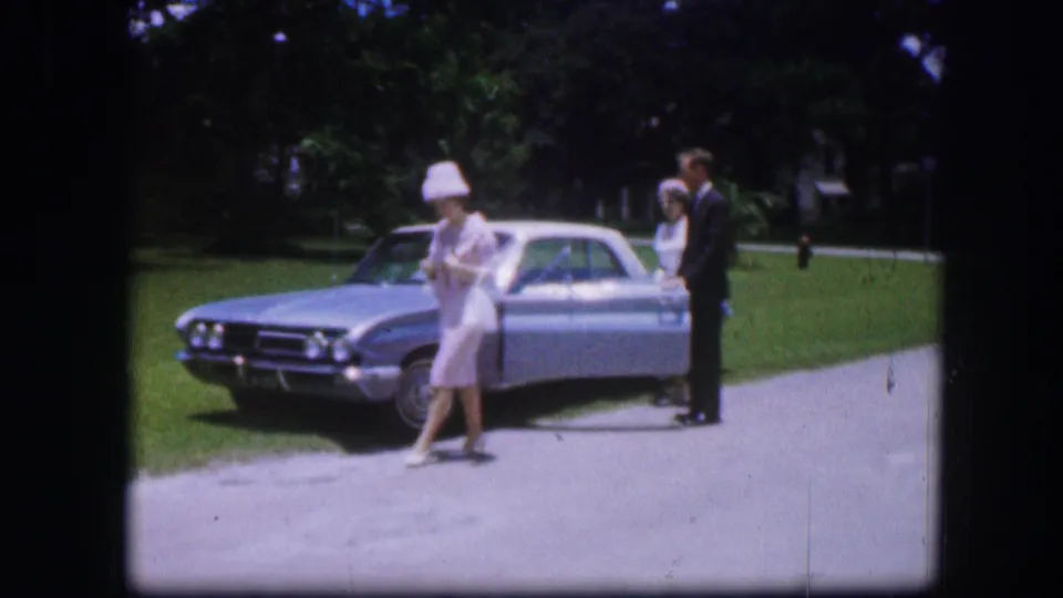 In 1967 Kirksville home-movie footage, a woman in a white hat stands near the front of a silver sedan with two adults beside the car.