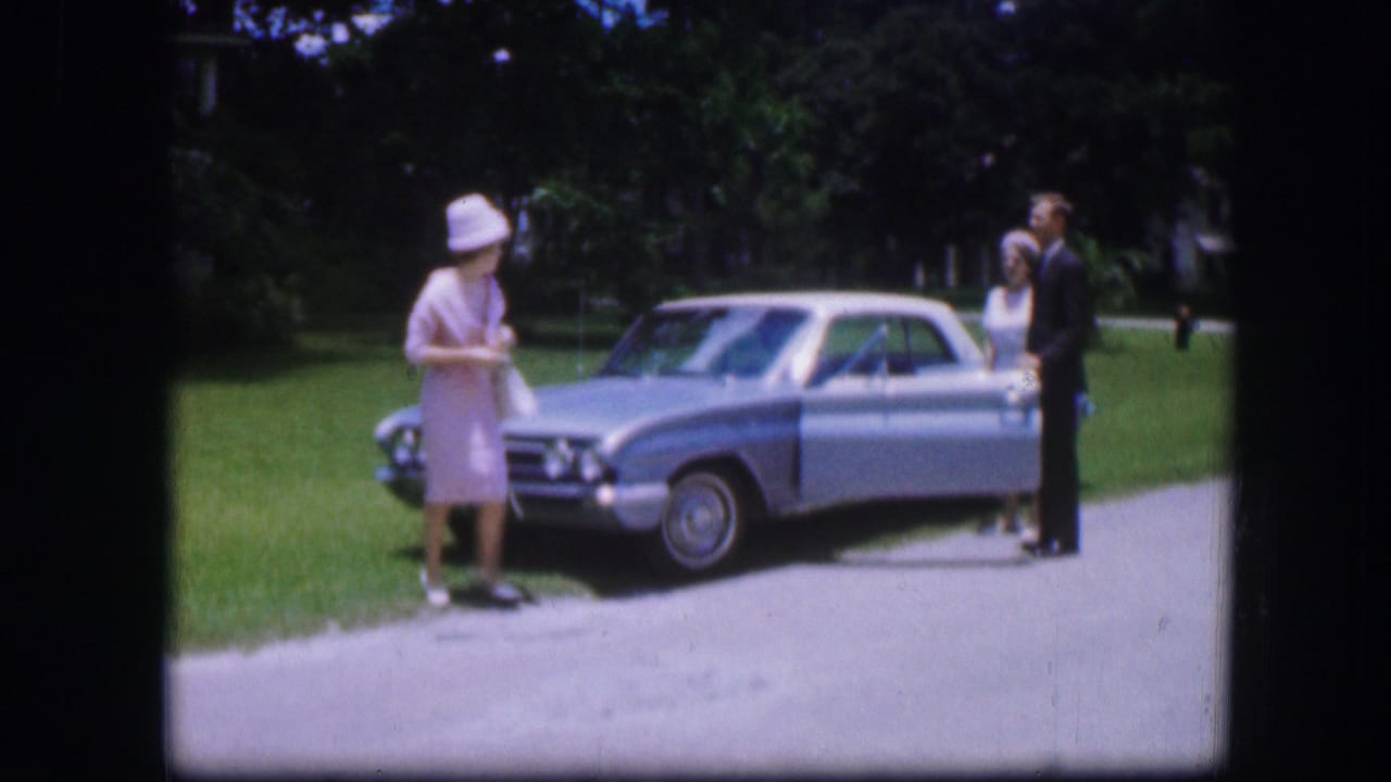 Woman in a pale dress stands in front of a silver sedan as two adults pose near the rear passenger side.