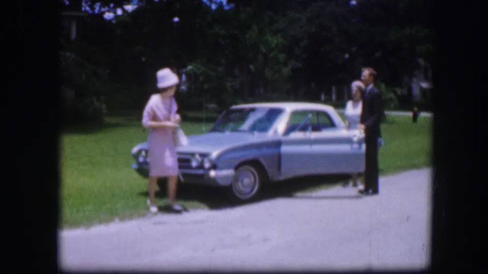 A woman in a pale outfit stands by the front fender of a silver sedan while a man and another woman pose nearby in Kirksville, Missouri.