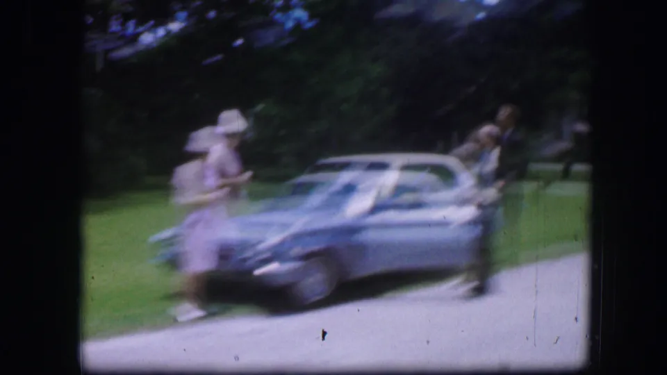 Motion blur from the 8mm camera captures a woman crossing near the silver sedan as the other adults remain by the car.