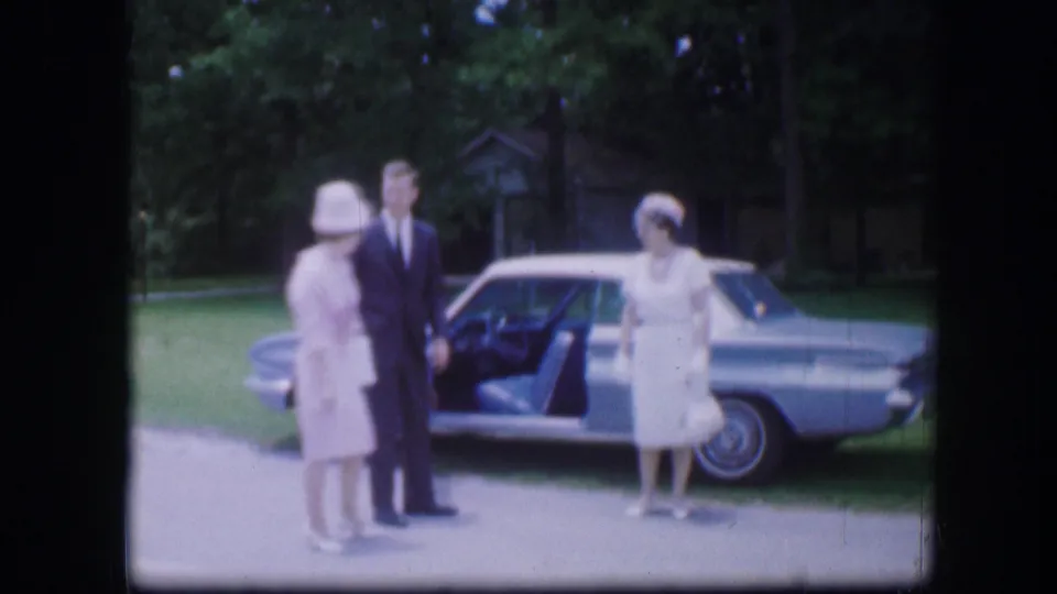 Two women in light dresses and hats and a man in a dark suit pose beside a silver sedan with an open door in 1967 Kirksville footage.