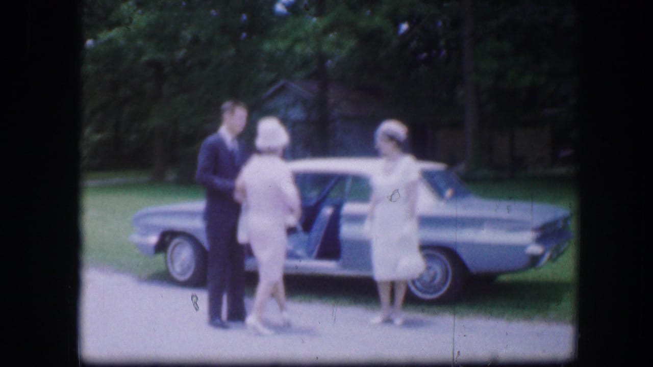 1967 Kirksville, Missouri home-movie frame showing a suited man beside a blue sedan with two women in white dresses and hats.