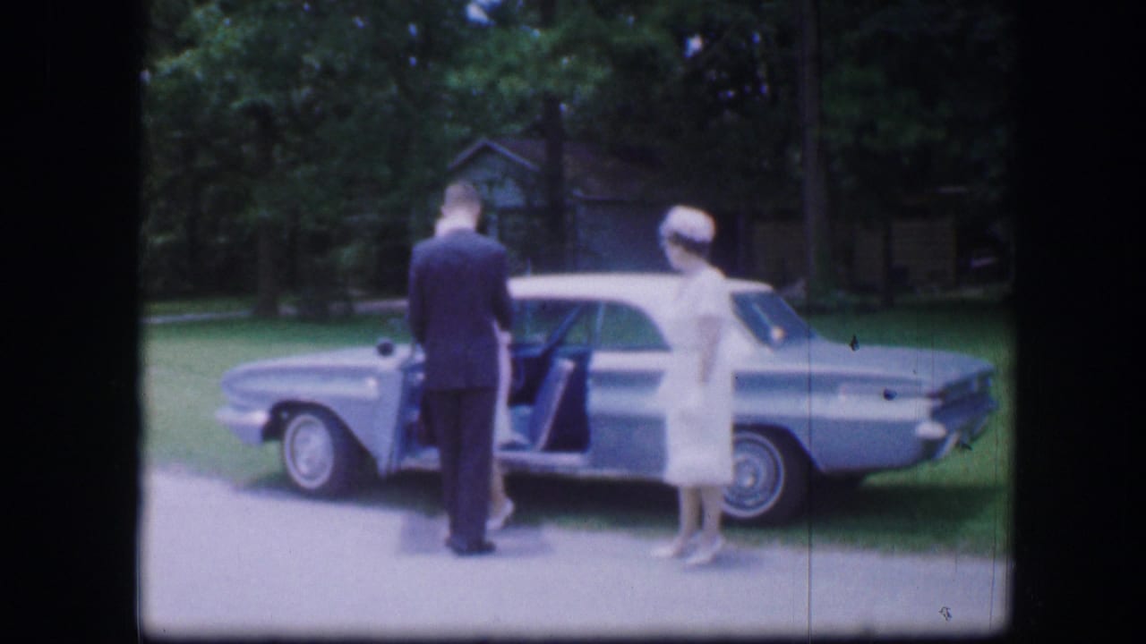 Man faces the open car door while two women in white stand beside the blue sedan.