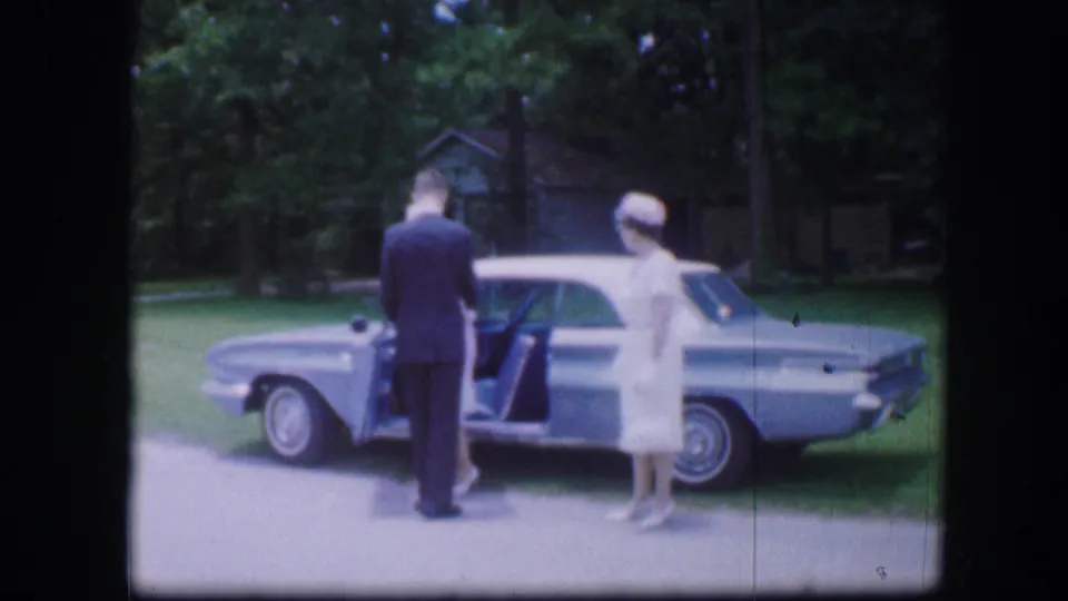 1967 Kirksville, Missouri frame with a man at an open blue sedan door and two women in white formal clothing nearby.