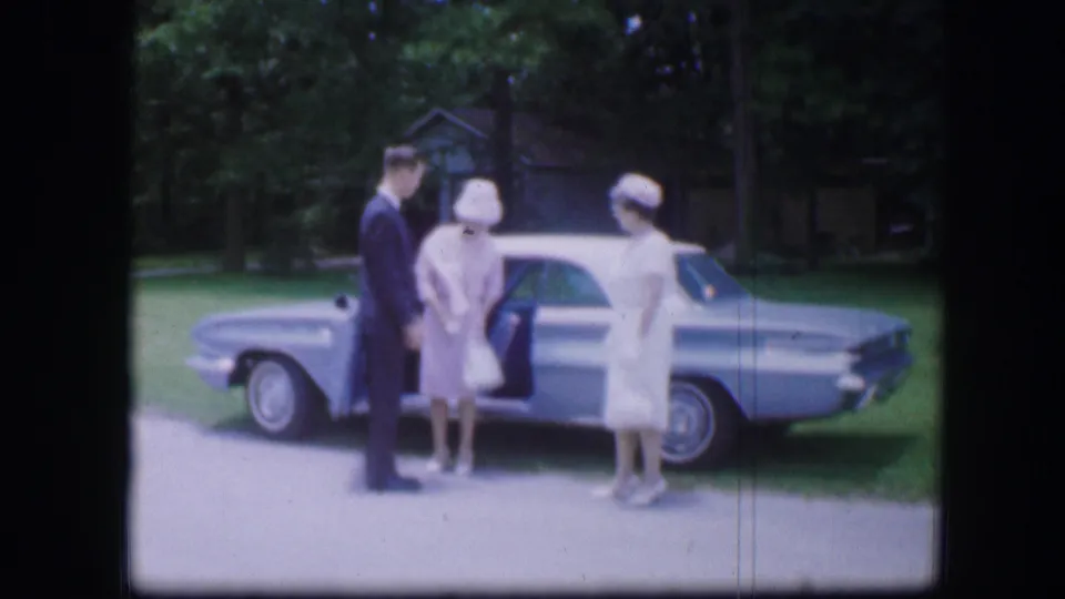 1967 Kirksville, Missouri home-movie frame showing a suited man and two women in white beside an open blue sedan door.