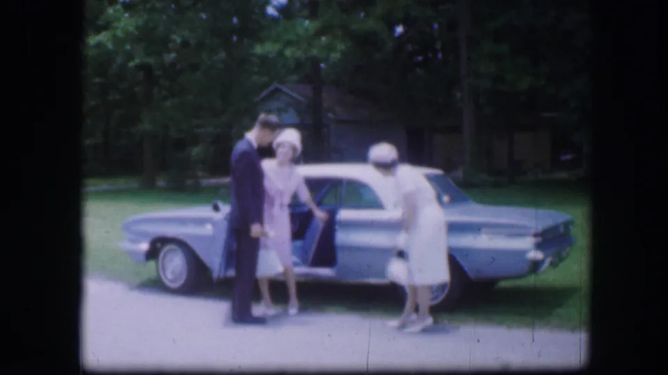1967 Kirksville, Missouri frame showing one woman leaning toward a blue sedan while a man and another woman stand beside her.
