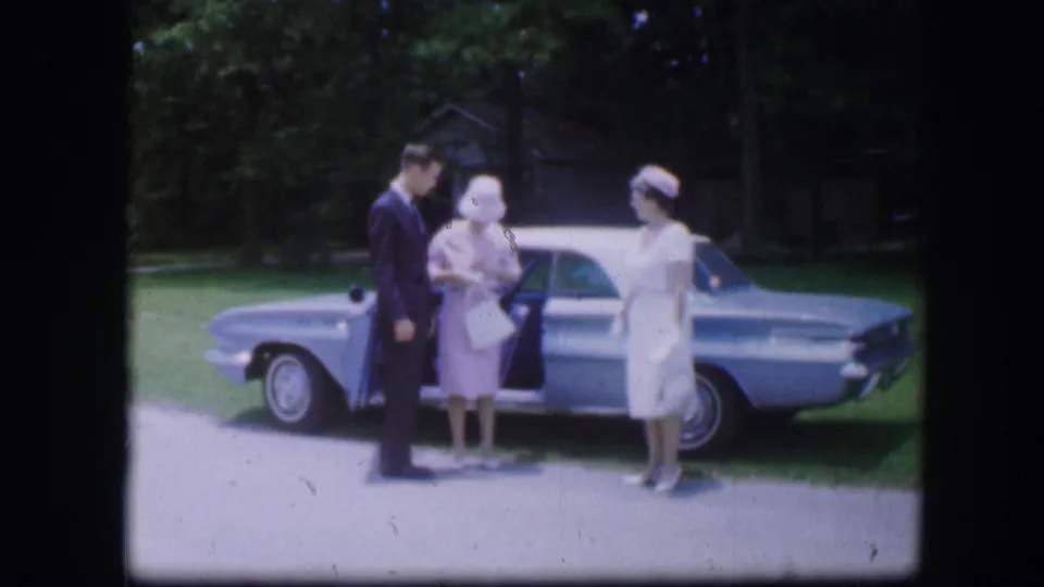 A suited man and two women in light clothing stand beside a parked car in 1967 Kirksville home-movie footage.