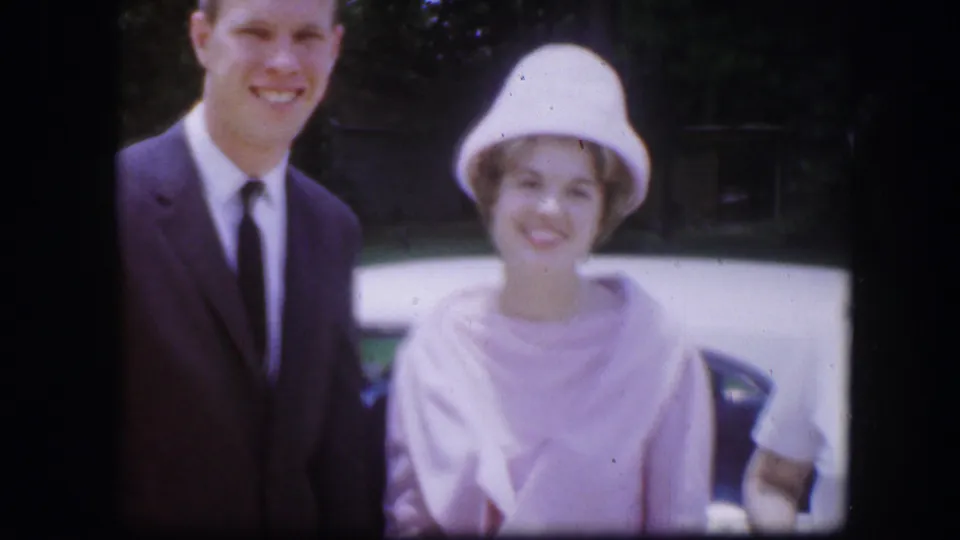 A man and woman smile toward the camera with a light car behind them in 1967 Kirksville home-movie footage.