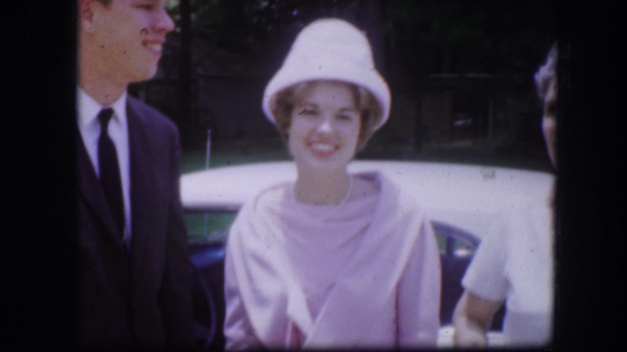 Adults standing near a blue car, including a smiling woman in a pale hat and pink wrap.