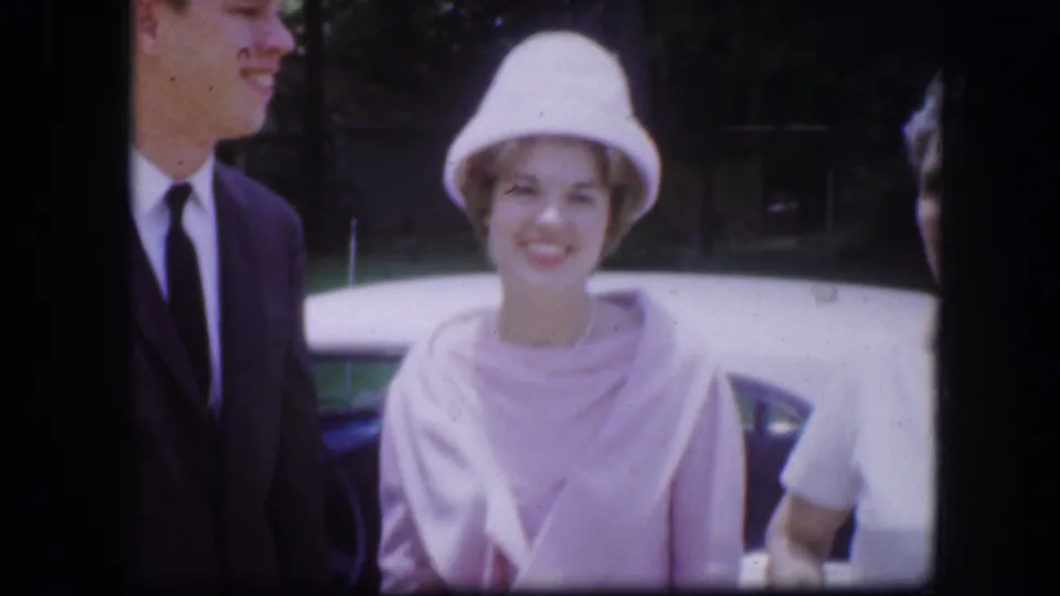1967 Kirksville home-movie frame showing adults posed beside a blue car, with a woman in a pale hat and pink wrap at center.