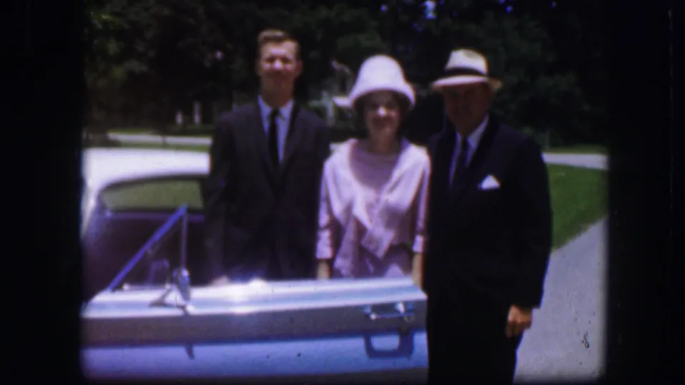 1967 Kirksville home-movie frame showing three adults formally dressed behind a blue car on a sunny day.