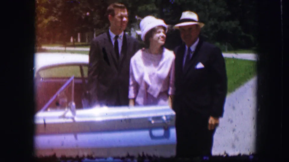 1967 Kirksville frame of three adults behind a blue car, with formal clothing, hats, pavement, and lawn visible.
