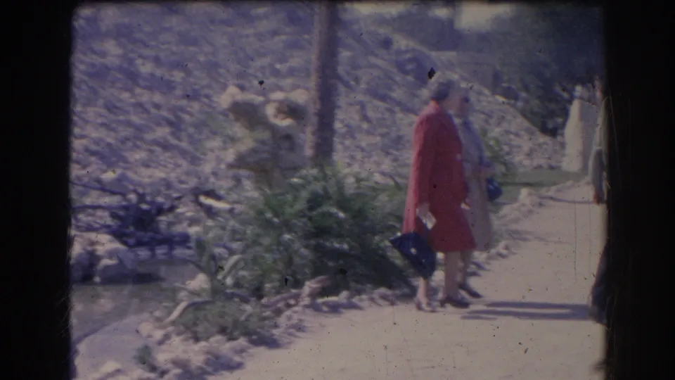 1969 Sarasota home-movie frame showing adults walking along a sunlit sidewalk near greenery and a low stone wall.
