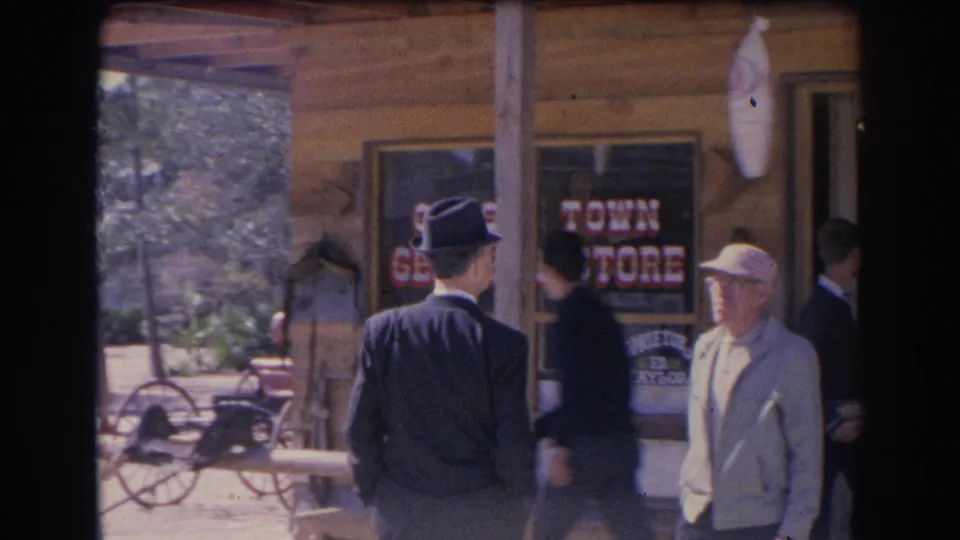 1969 Sarasota frame of men near a wooden storefront porch with painted general store lettering visible in the windows.