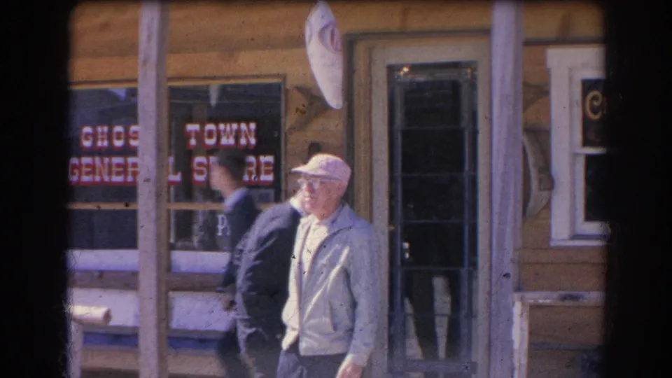 A man in a light jacket and cap stands near a wooden Sarasota storefront with general store window lettering in this 1969 home-movie frame.