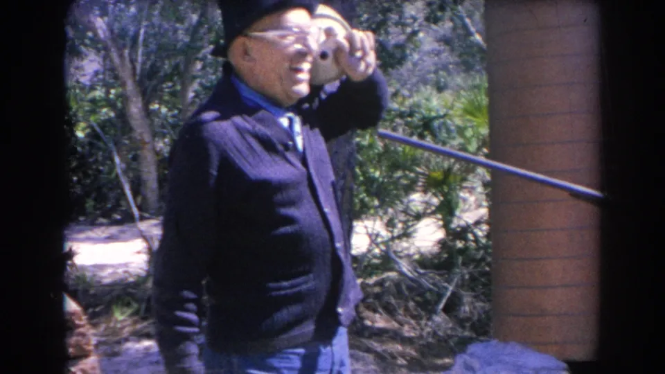 The 1969 Sarasota footage shows an older man in a dark cardigan and hat pausing outside near greenery while shielding his eyes.