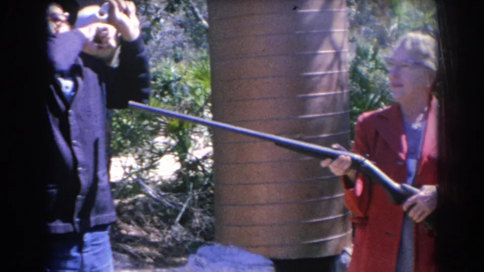 A woman in a red coat holds a long pole or gun-like object beside a wooden wall in the 1969 Sarasota home-movie scene.