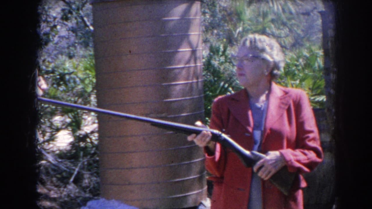 Woman in a red jacket holding a long gun outdoors beside a brick structure.